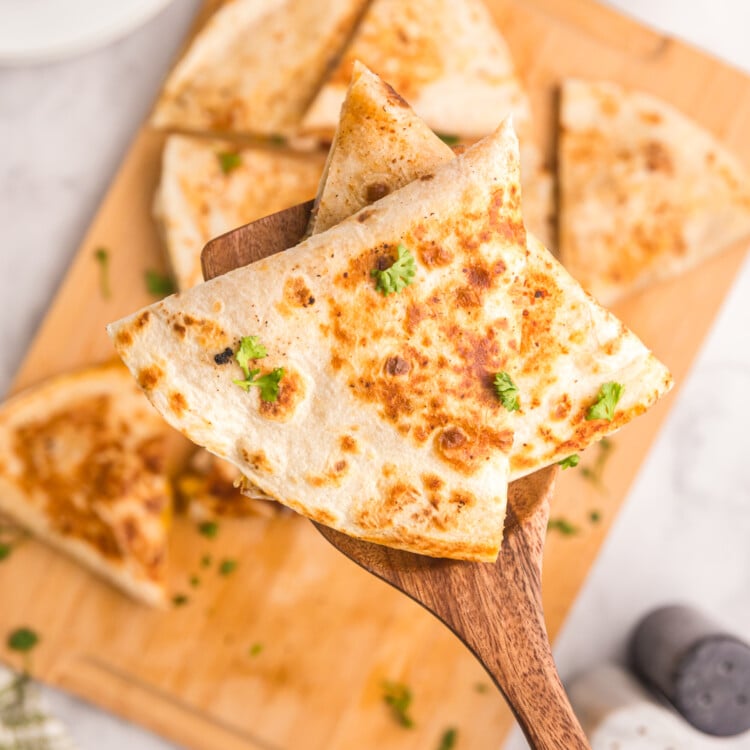 Spatula holding a wedge of browned quesadilla garnished with fresh parsley over a cutting board.