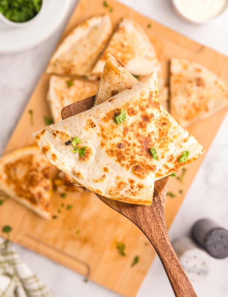 Spatula holding a wedge of browned quesadilla garnished with fresh parsley over a cutting board.