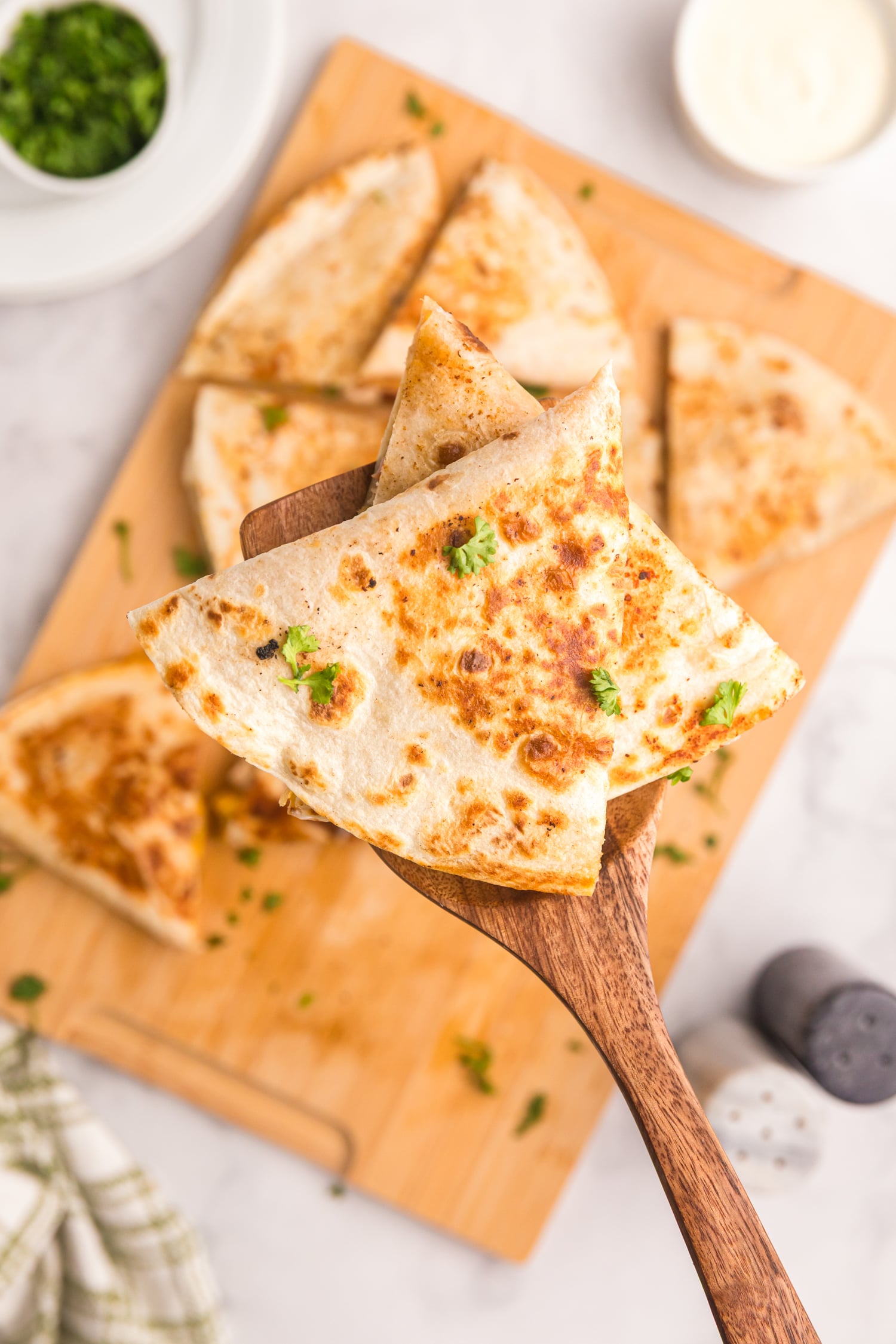 Spatula holding a wedge of browned quesadilla garnished with fresh parsley over a cutting board.