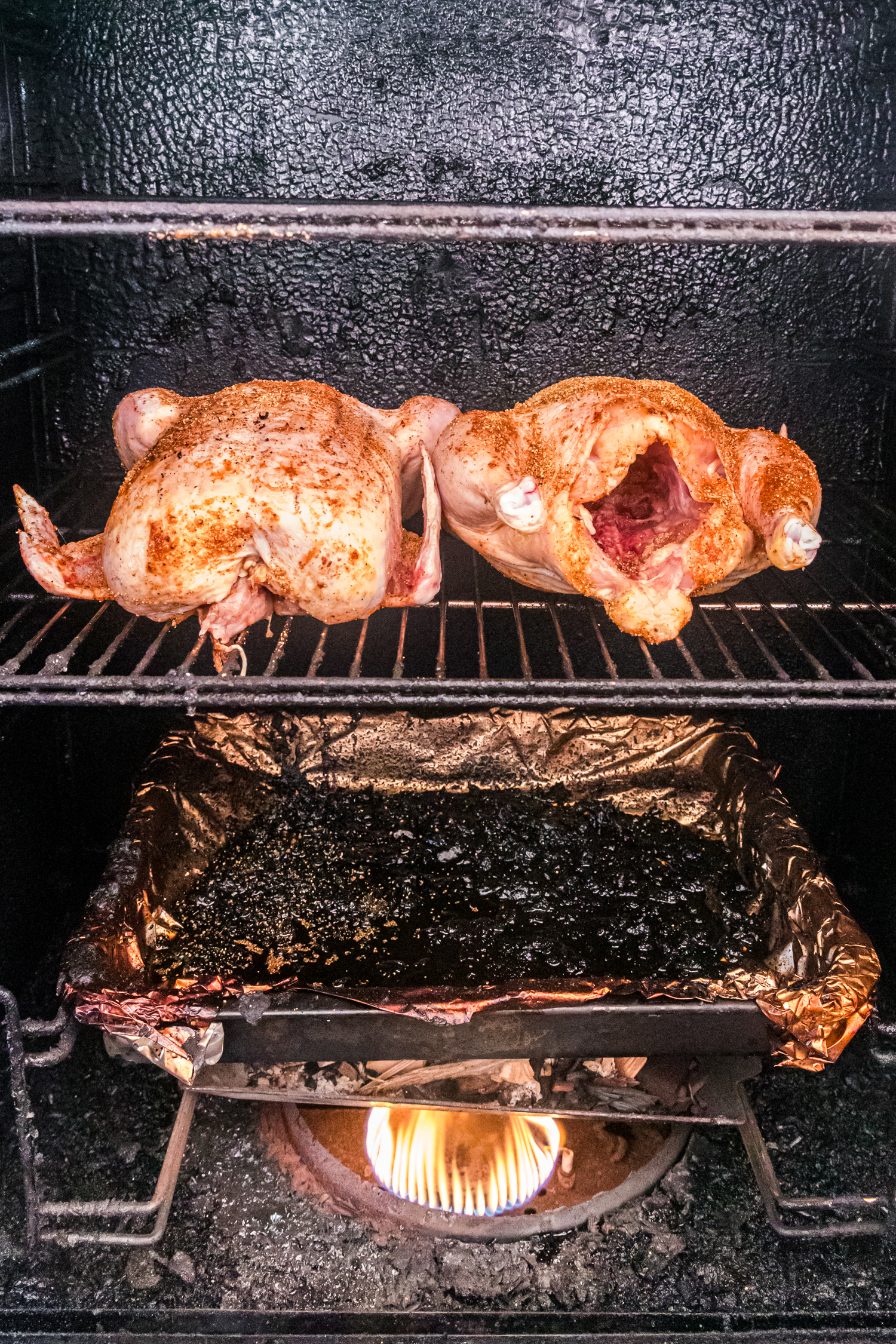 Two seasoned whole chickens placed on a smoker rack above a drip pan with fire burning below.