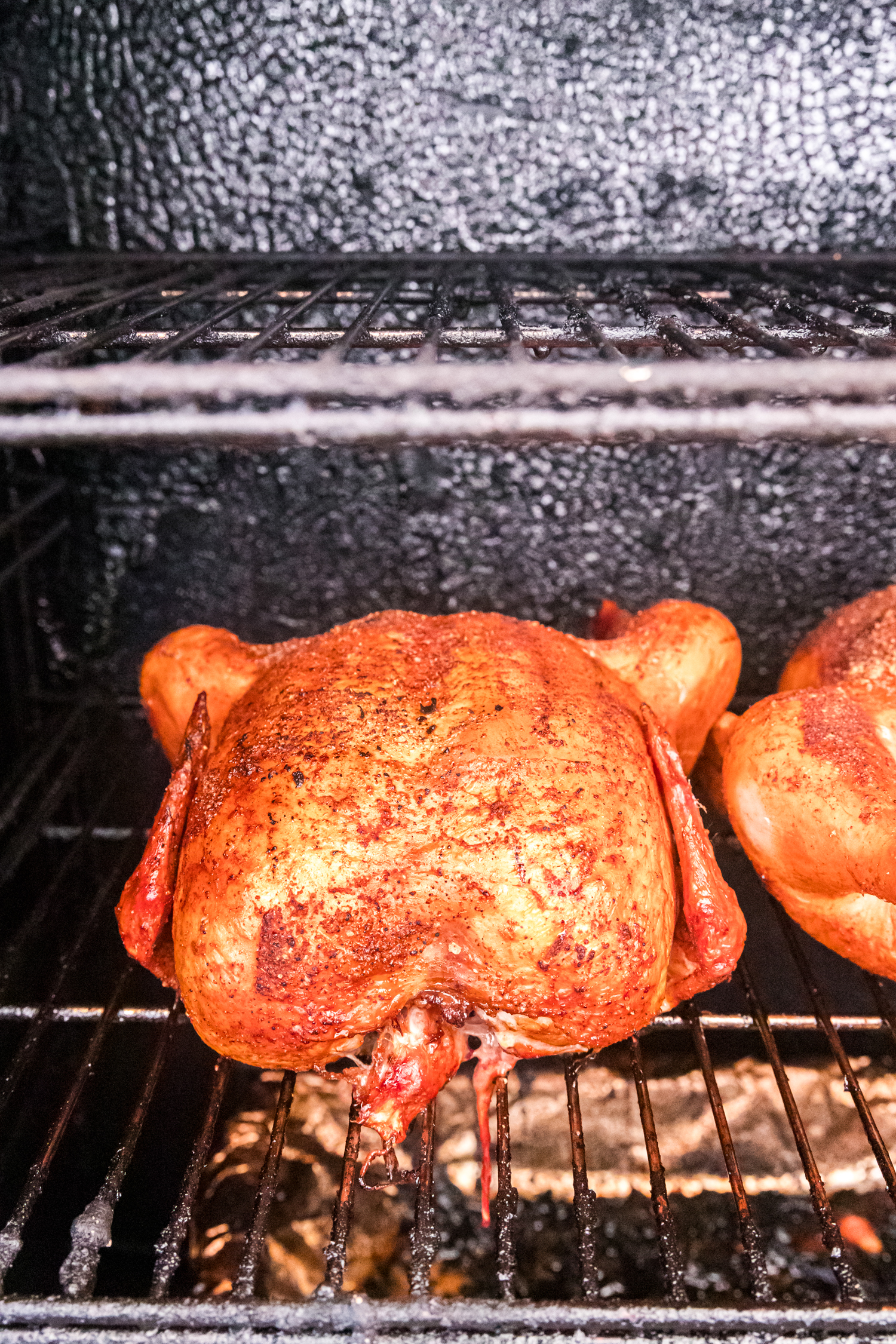 Whole chicken smoking on a grill rack, golden brown with BBQ seasoning and crispy skin inside a smoker.
