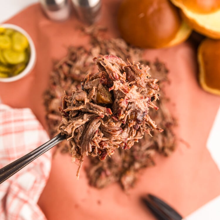 Close-up of a fork lifting a portion of shredded smoked chuck roast, with buns and pickles blurred in the background.