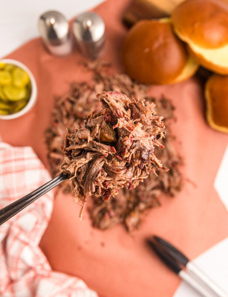 Close-up of a fork lifting a portion of shredded smoked chuck roast, with buns and pickles blurred in the background.