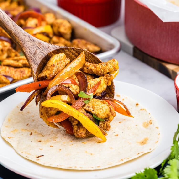Wooden spoon placing cooked chicken, peppers, and onions onto a warm flour tortilla.