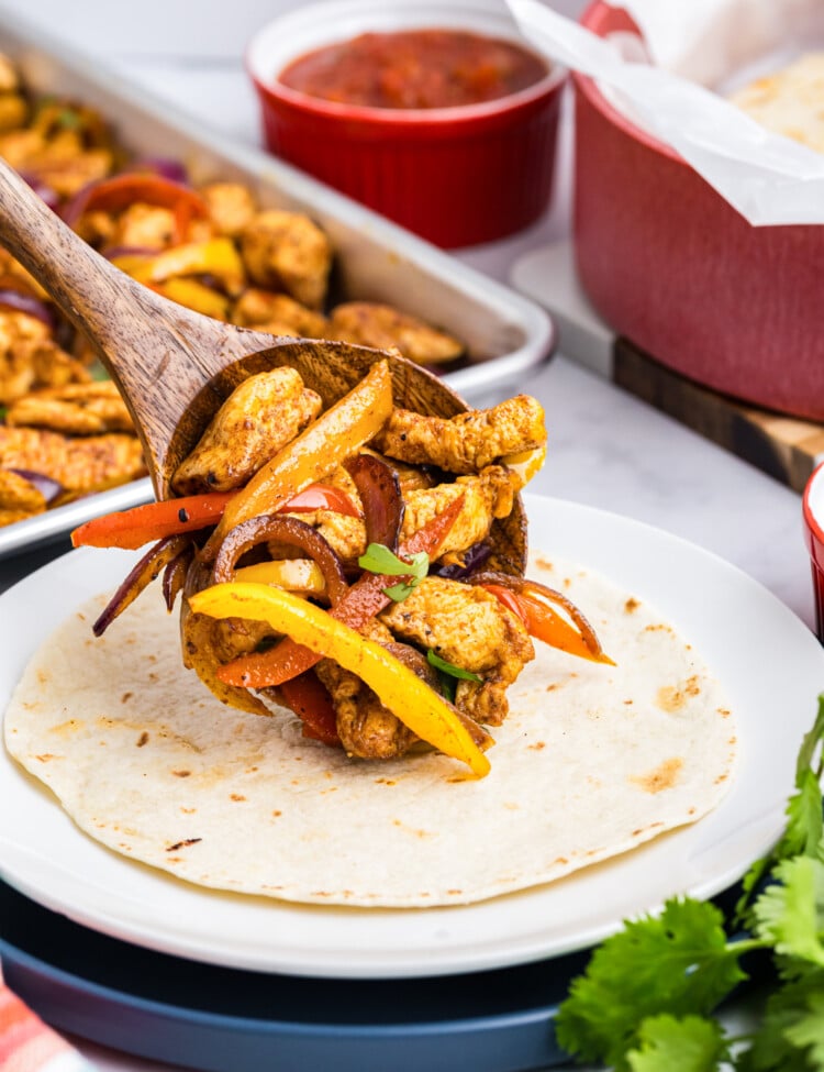 Wooden spoon placing cooked chicken, peppers, and onions onto a warm flour tortilla.