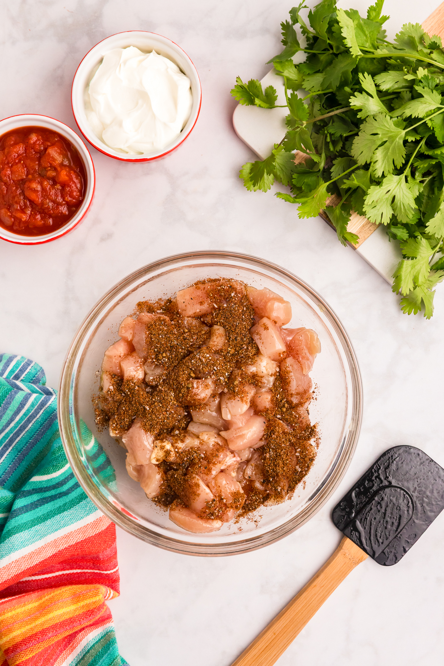 Raw chicken pieces coated with fajita seasoning in a glass bowl, with salsa, sour cream, and fresh cilantro nearby.