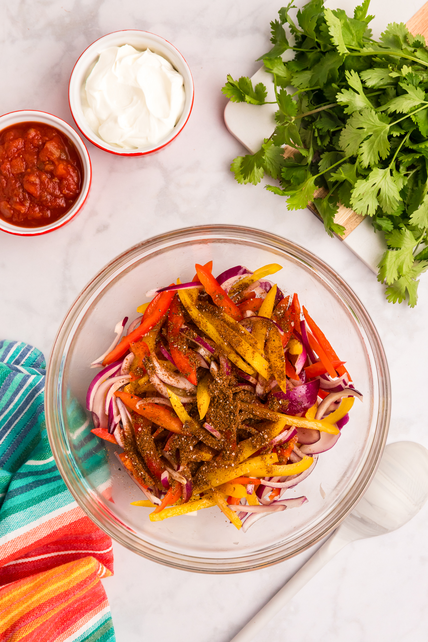 Sliced bell peppers and red onions tossed with seasoning in a glass bowl on a countertop.