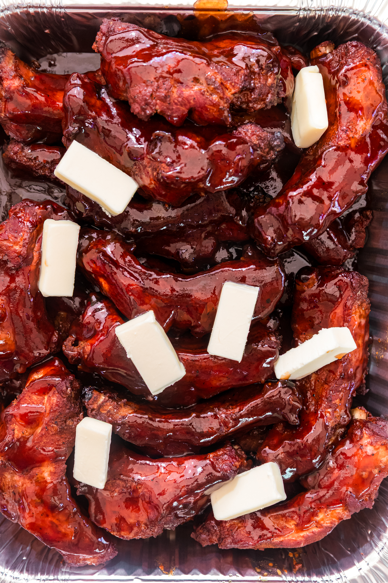 Smoked pork ribs coated in glossy barbecue sauce with slices of butter, resting in an aluminum pan.