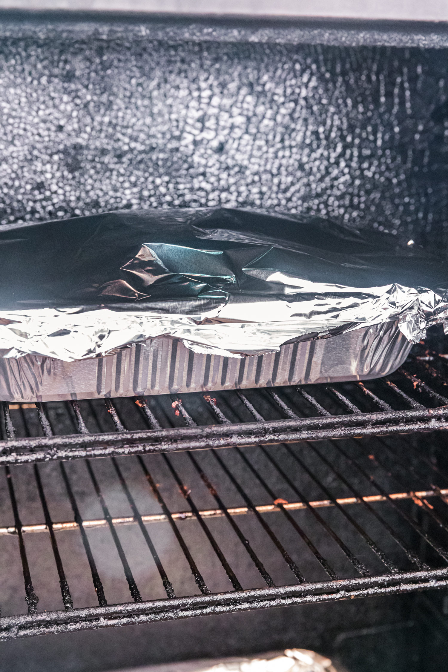Foil-covered aluminum pan of ribs placed inside a smoker on metal grates.