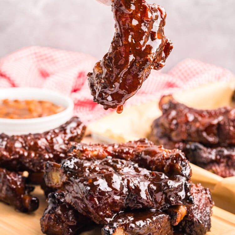A hand holding a single barbecue-glazed rib dripping sauce above a stack of ribs on a wooden board.