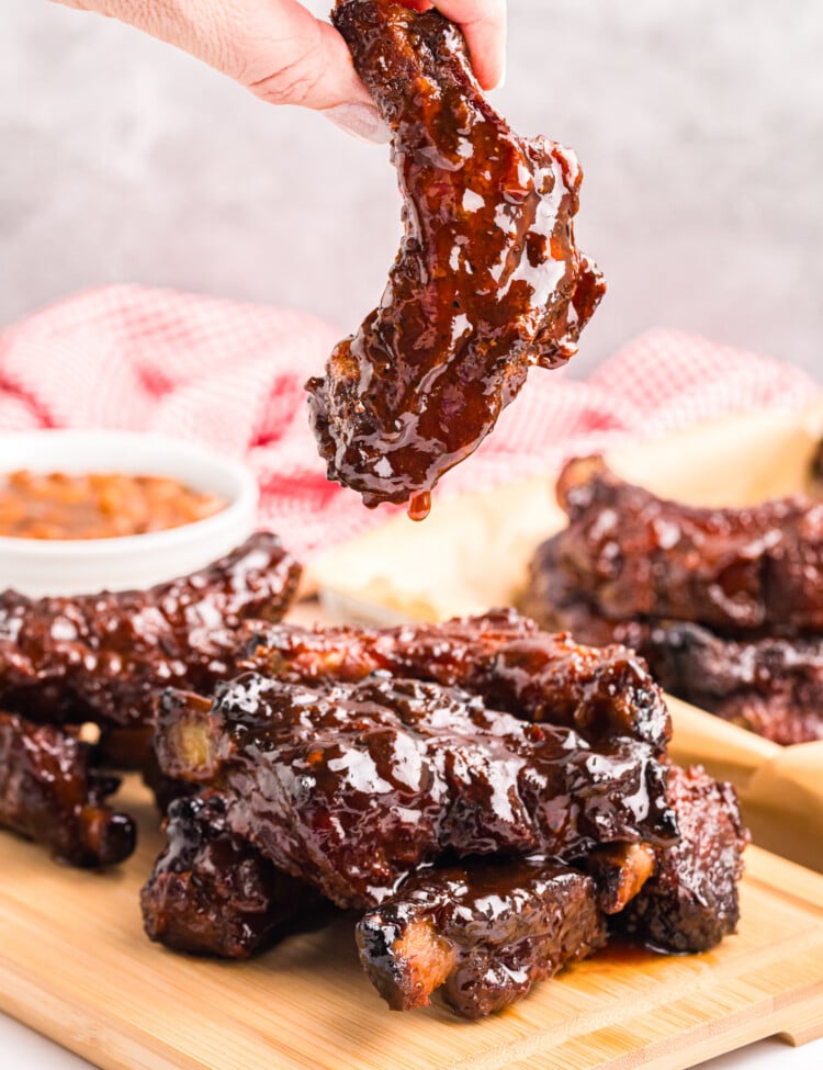 A hand holding a single barbecue-glazed rib dripping sauce above a stack of ribs on a wooden board.