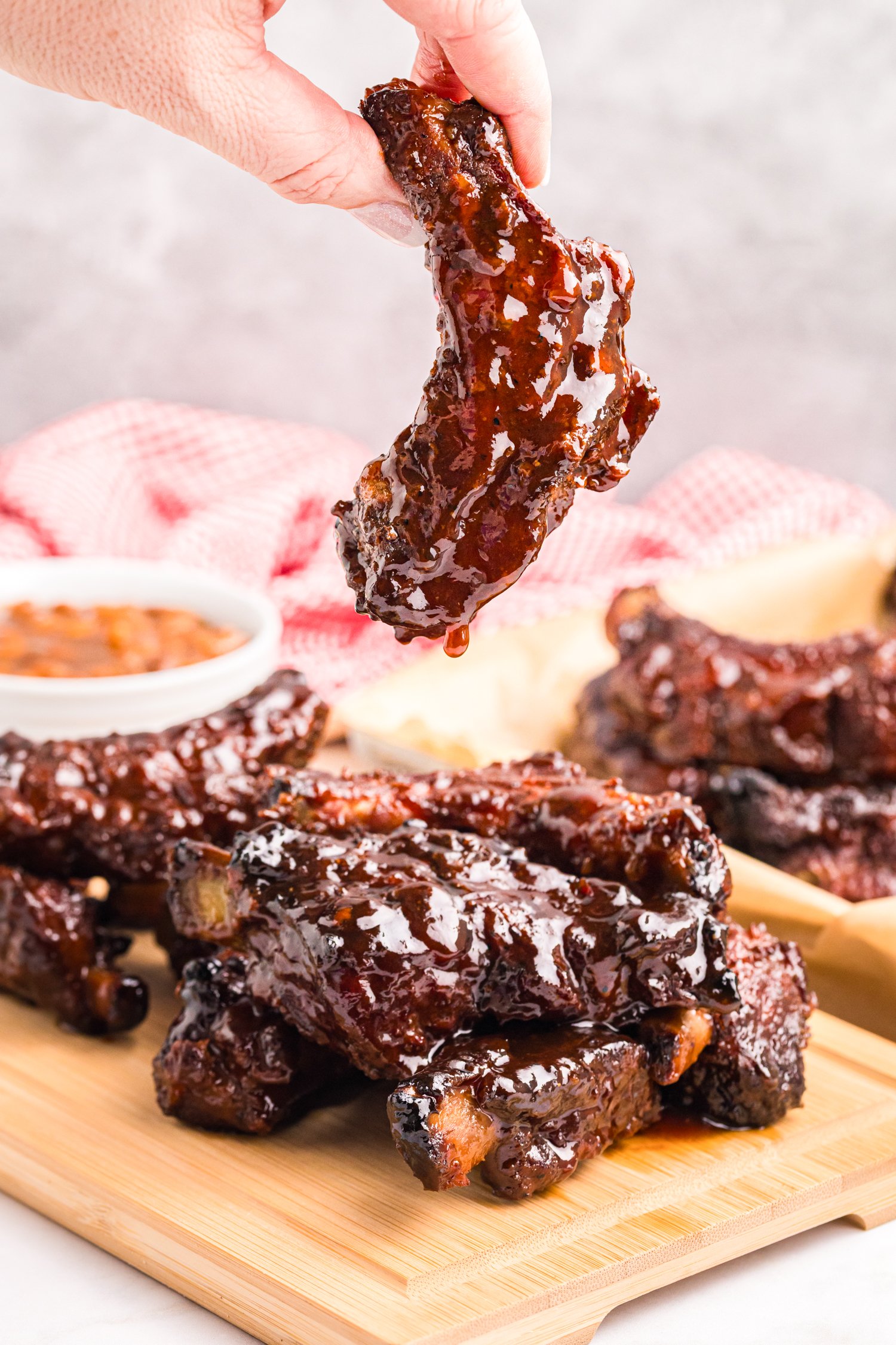 A hand holding a single barbecue-glazed rib dripping sauce above a stack of ribs on a wooden board.