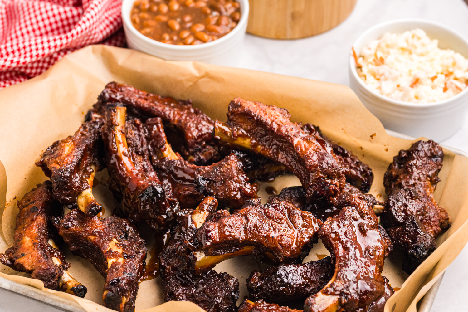 Pile of sticky barbecue pork ribs served on parchment paper with baked beans and coleslaw in the background.