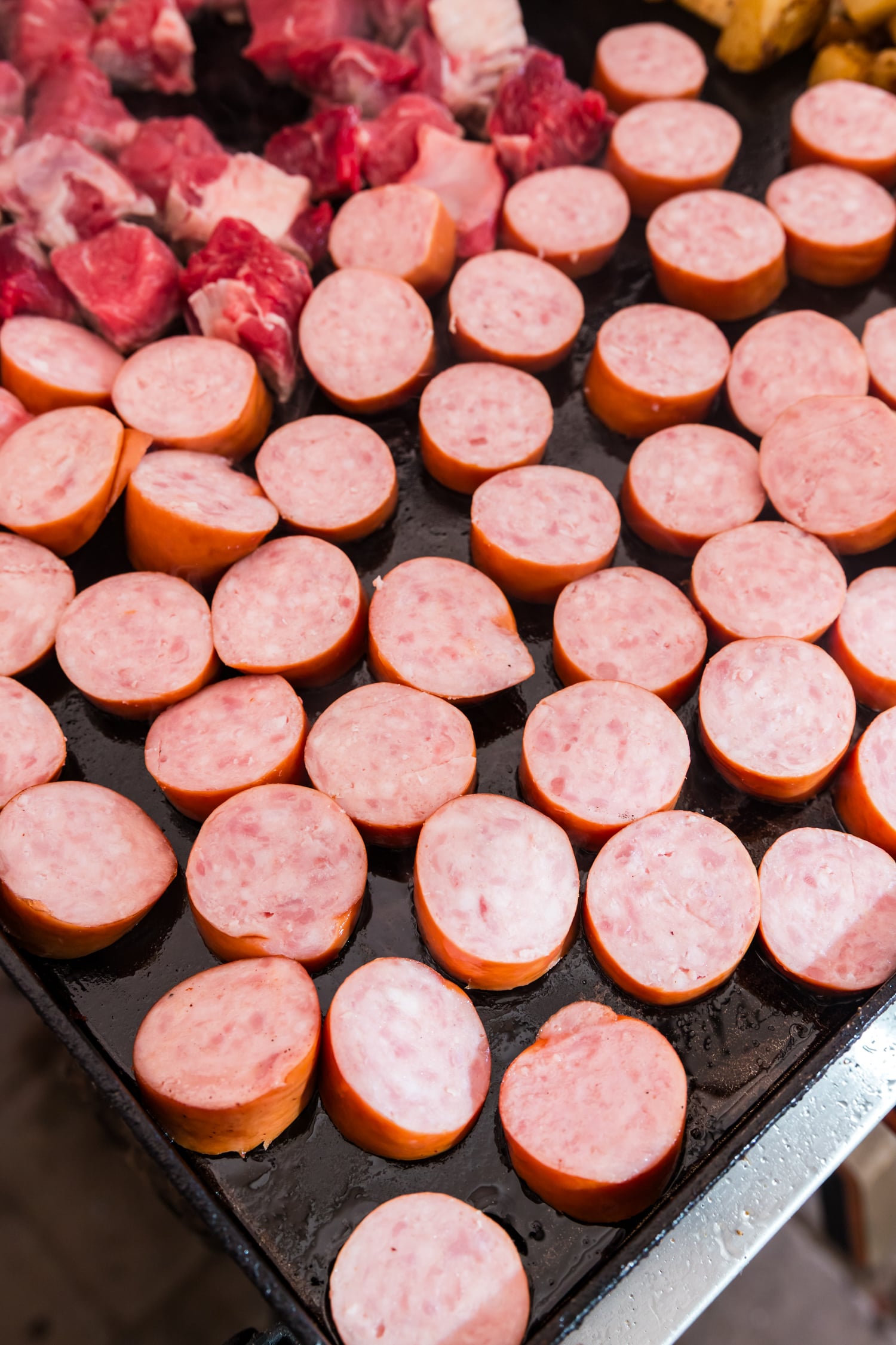 Sliced sausage rounds and chunks of raw beef browning on a flat-top griddle.
