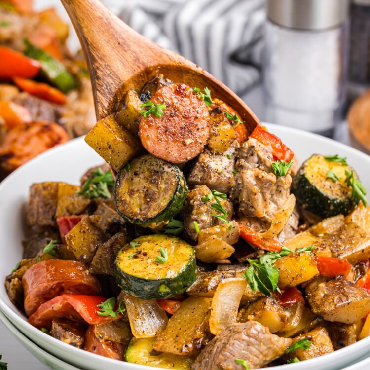 Wooden spoon lifting a serving of sausage, beef, potatoes, zucchini, and peppers from a bowl, garnished with chopped parsley.