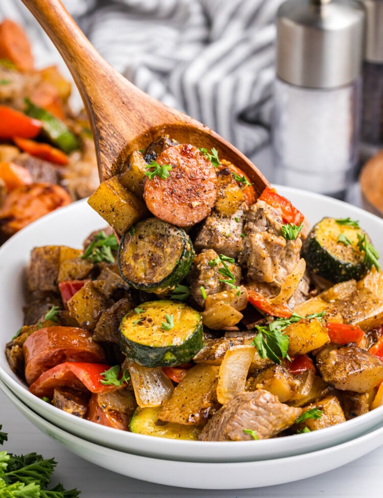 Wooden spoon lifting a serving of sausage, beef, potatoes, zucchini, and peppers from a bowl, garnished with chopped parsley.