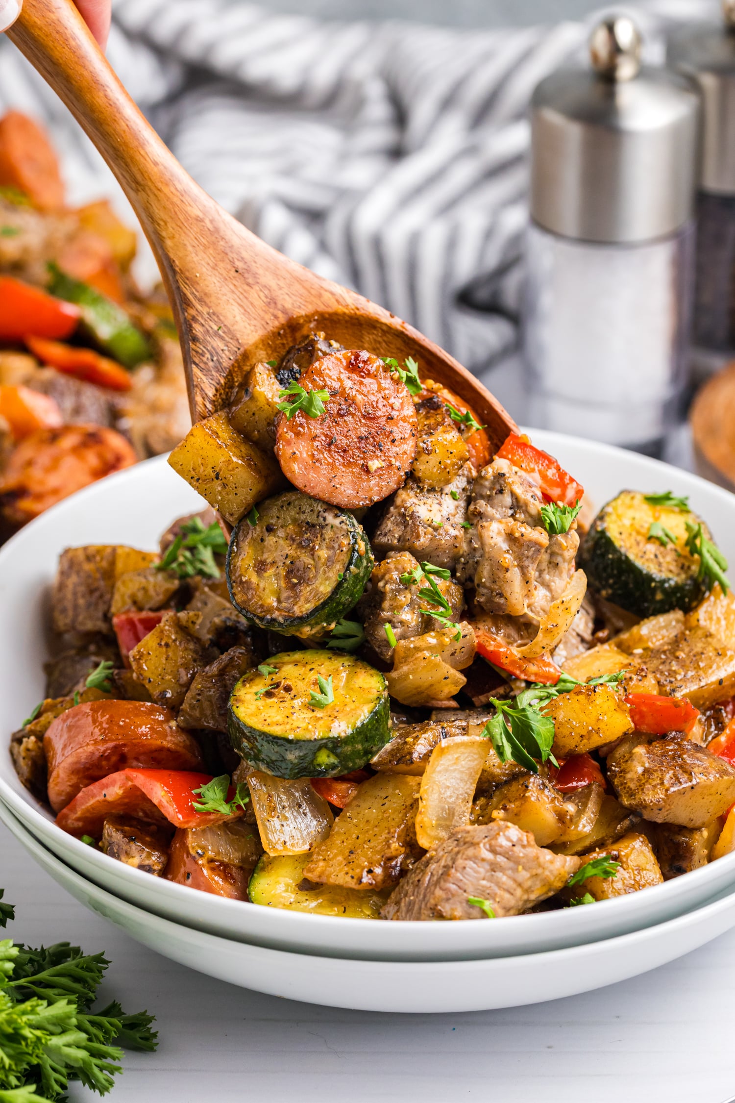 Wooden spoon lifting a serving of sausage, beef, potatoes, zucchini, and peppers from a bowl, garnished with chopped parsley.