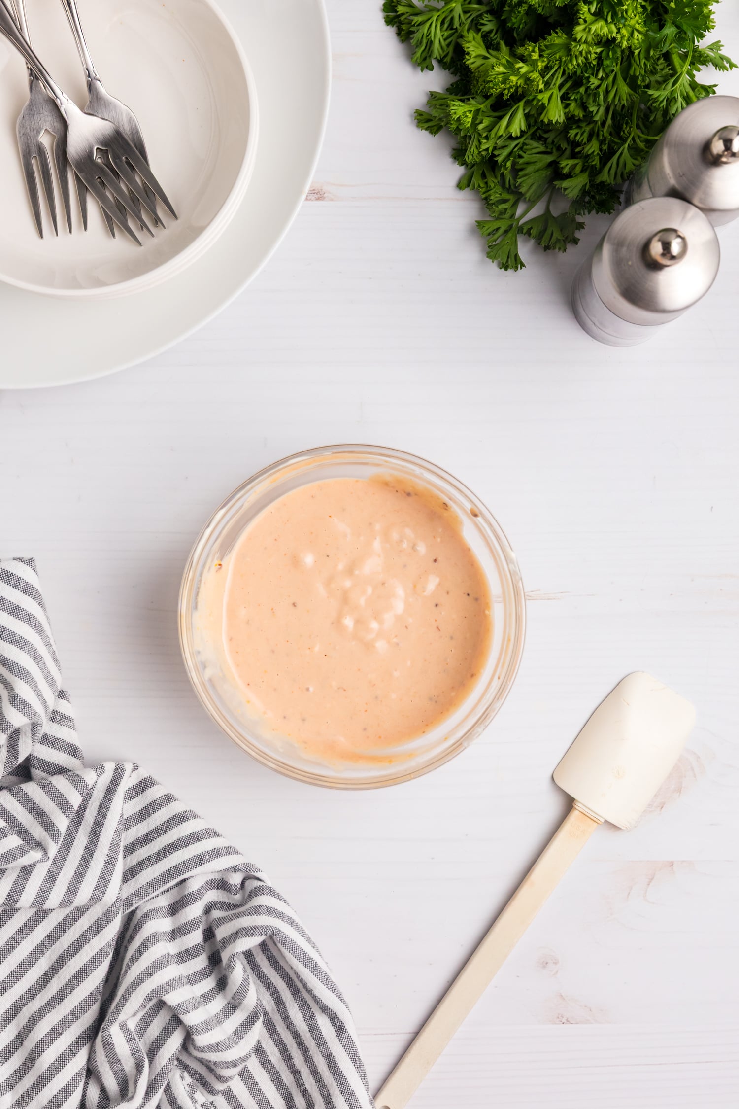 Small bowl of creamy pink sauce on a white surface with a striped towel, spatula, plates and forks, fresh parsley, and salt and pepper shakers nearby.