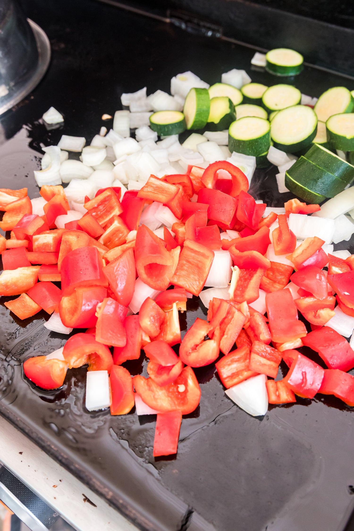 Chopped red bell pepper, diced onion, and sliced zucchini spread across an oiled flat-top griddle.