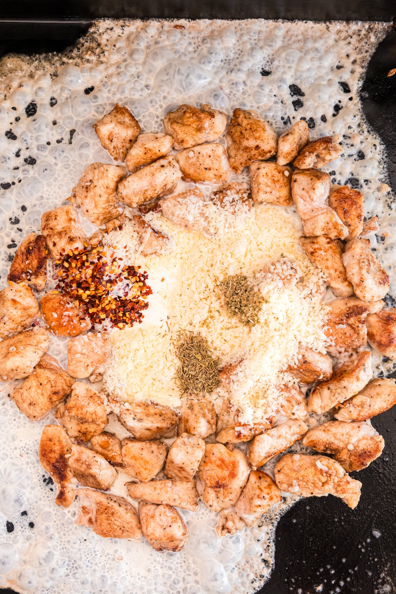 Cooked chicken pieces are arranged in a circle on a griddle while cream, grated cheese, red pepper flakes, and dried herbs melt together in the center to form a sauce.