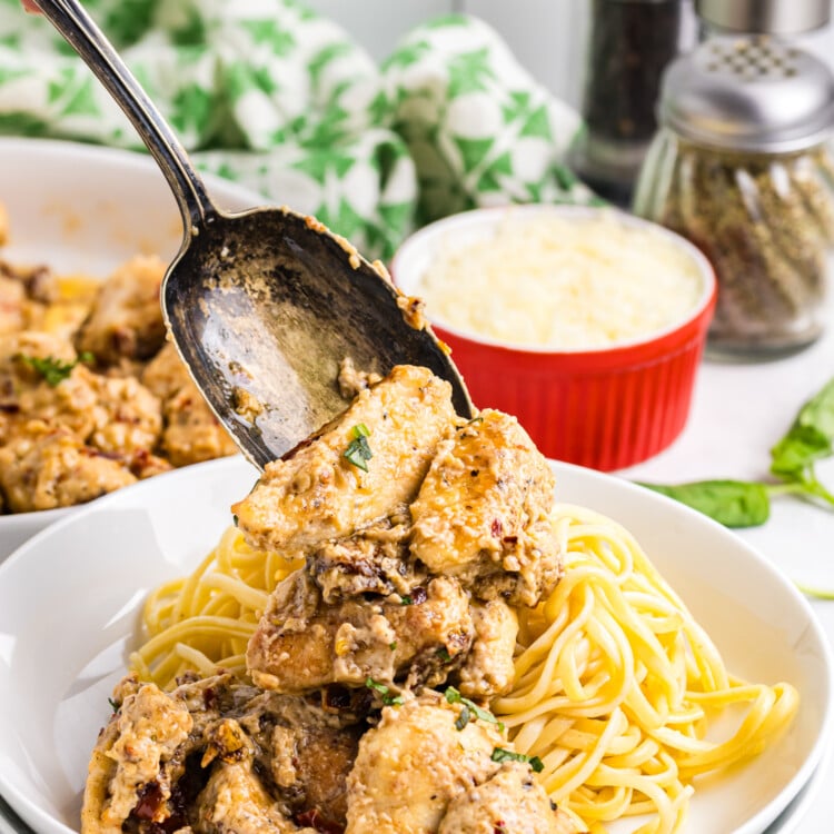 A spoon lifts creamy sun-dried tomato chicken over a bowl of cooked pasta, with grated cheese and seasonings visible in the background.