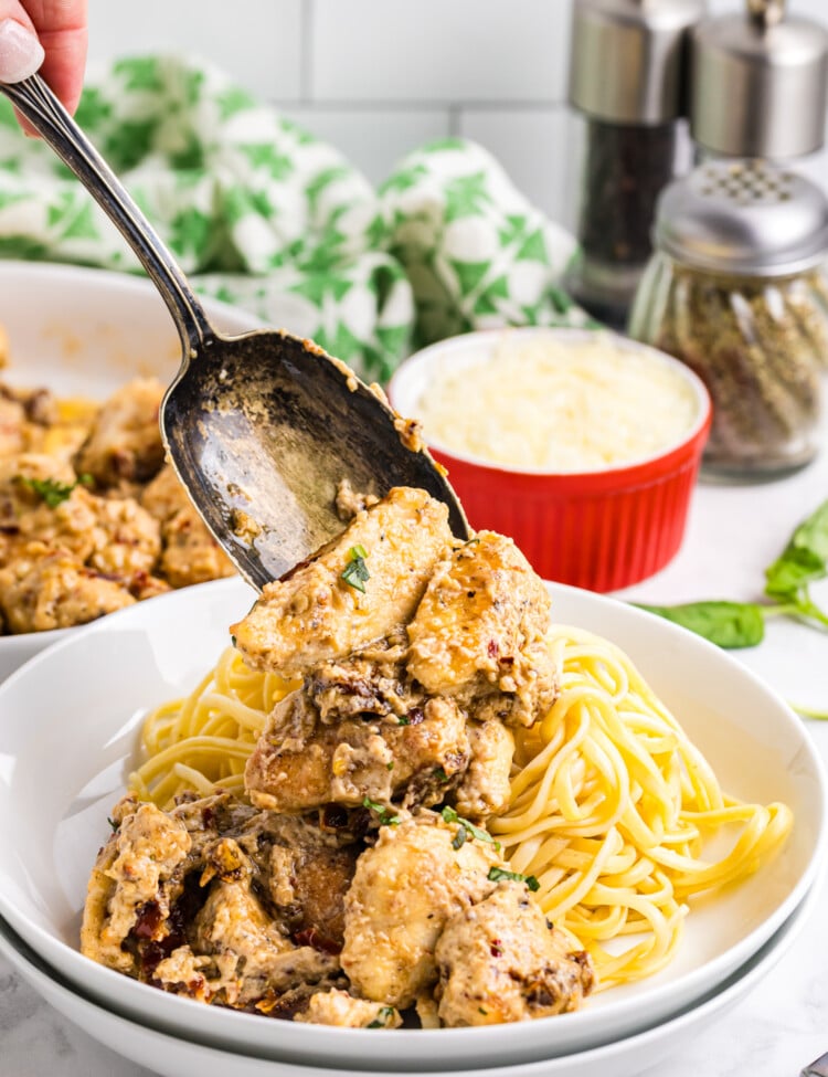 A spoon lifts creamy sun-dried tomato chicken over a bowl of cooked pasta, with grated cheese and seasonings visible in the background.