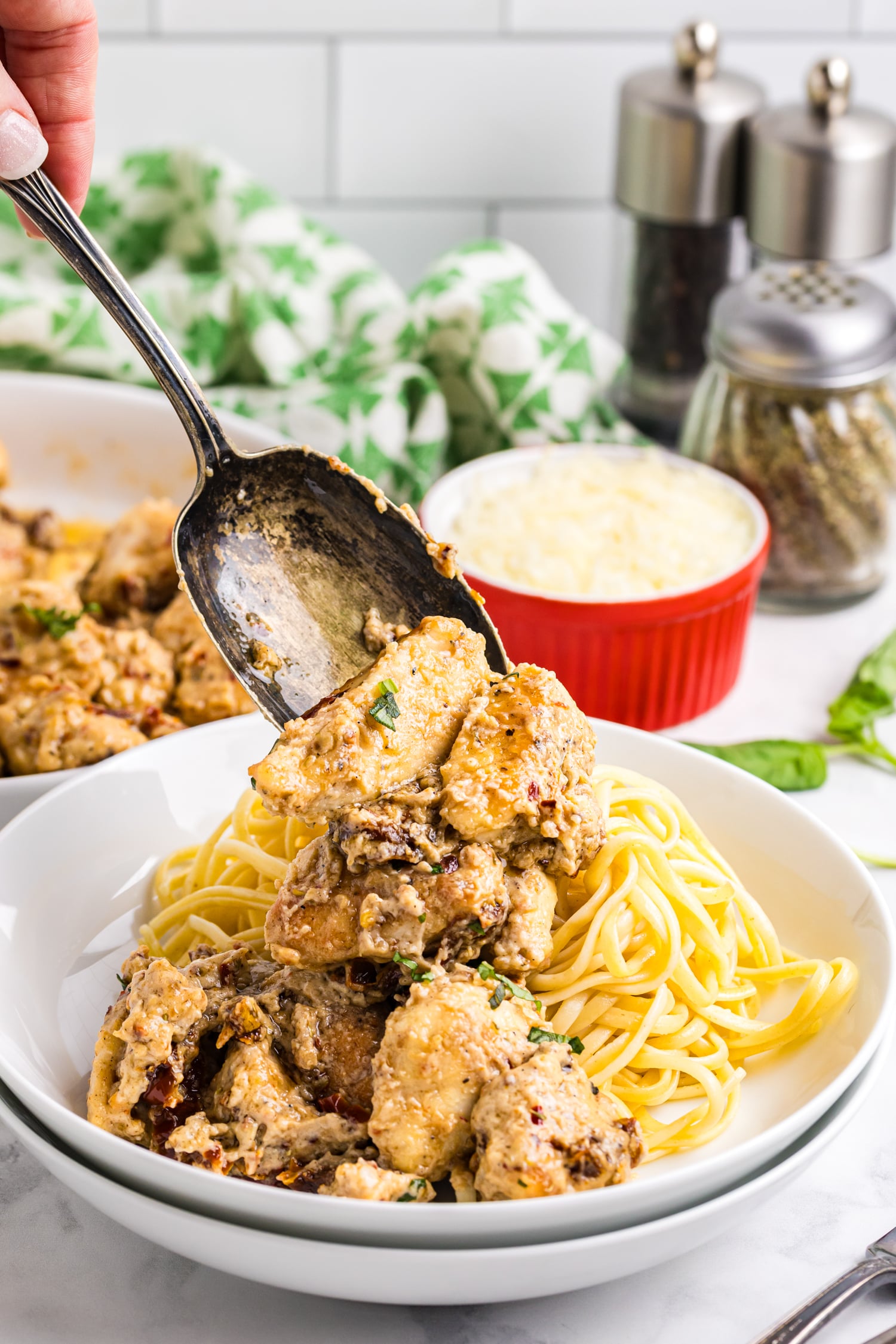 A spoon lifts creamy sun-dried tomato chicken over a bowl of cooked pasta, with grated cheese and seasonings visible in the background.