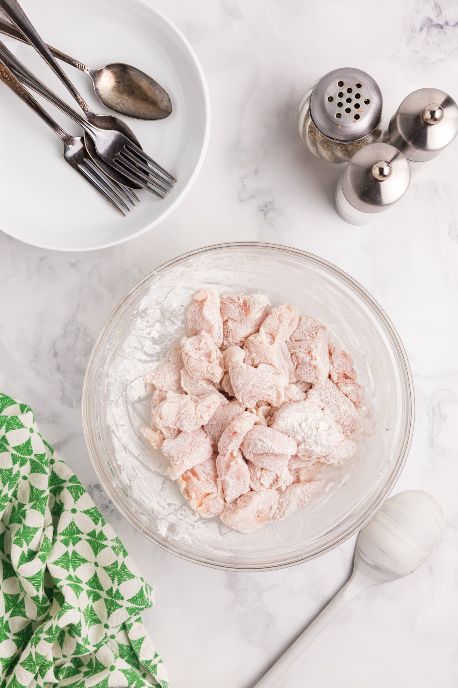 Raw chicken pieces coated in flour sit in a glass bowl on a marble countertop, surrounded by a white plate, utensils, salt and pepper shakers, and a green patterned kitchen towel.