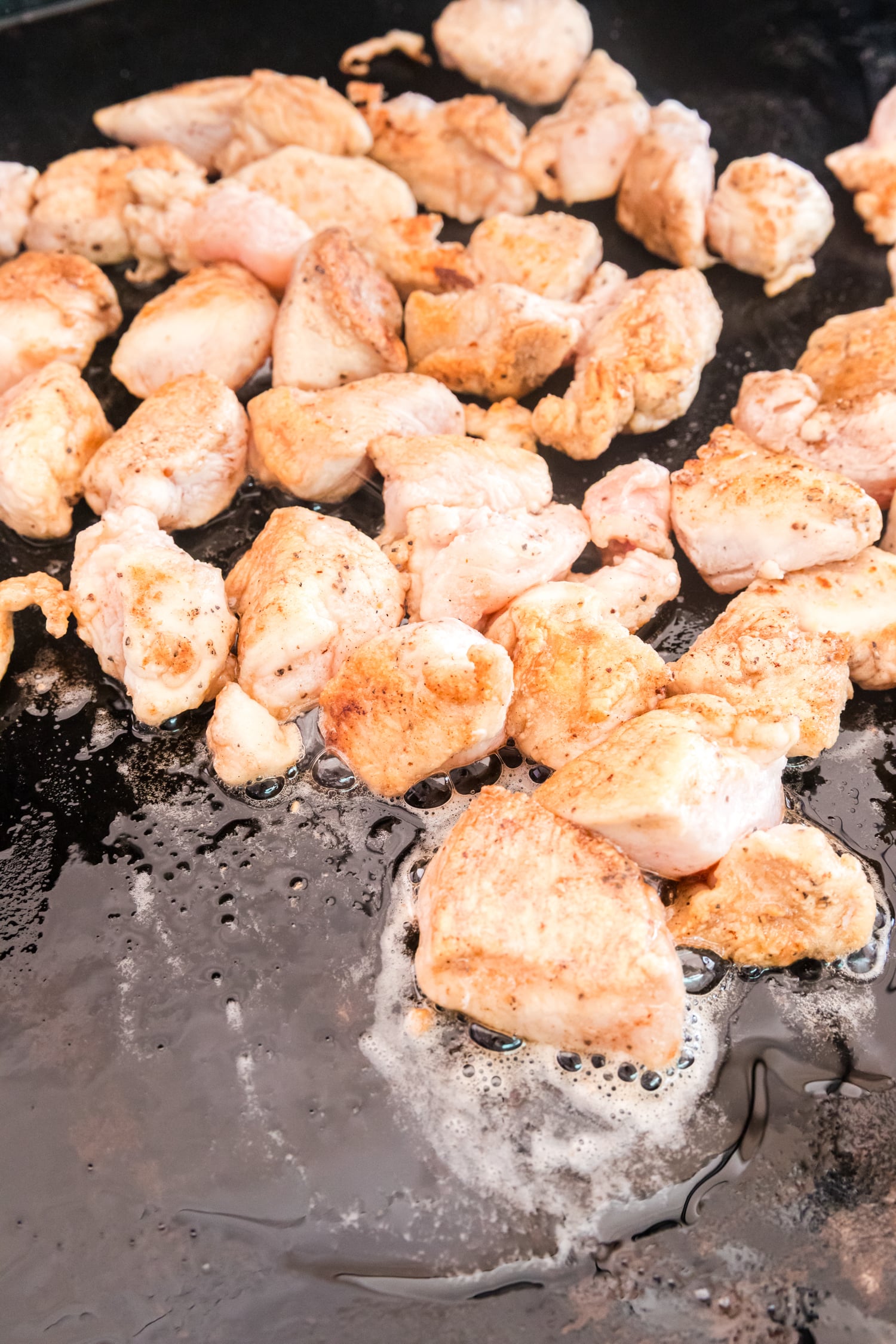 Bite-sized chicken pieces cook on a flat-top griddle, lightly browned on the outside with oil bubbling around them.