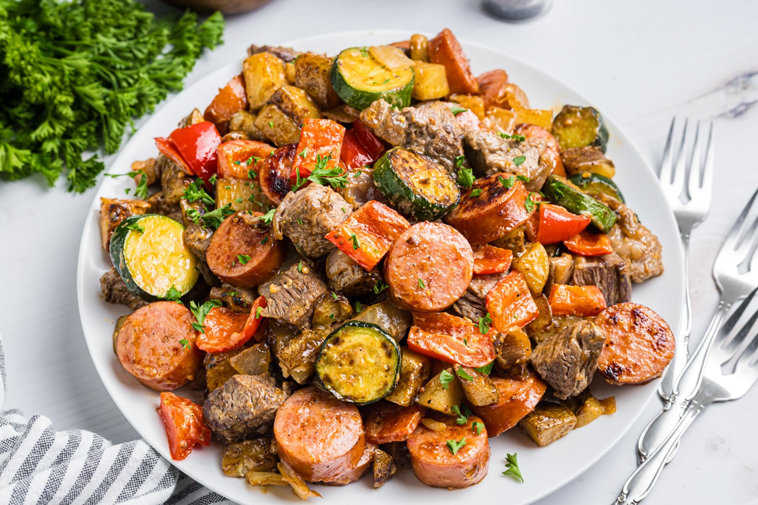 Platter piled with cowboy stir-fry—sausage, beef, potatoes, zucchini, peppers, and onions—sprinkled with parsley, with forks on the side.