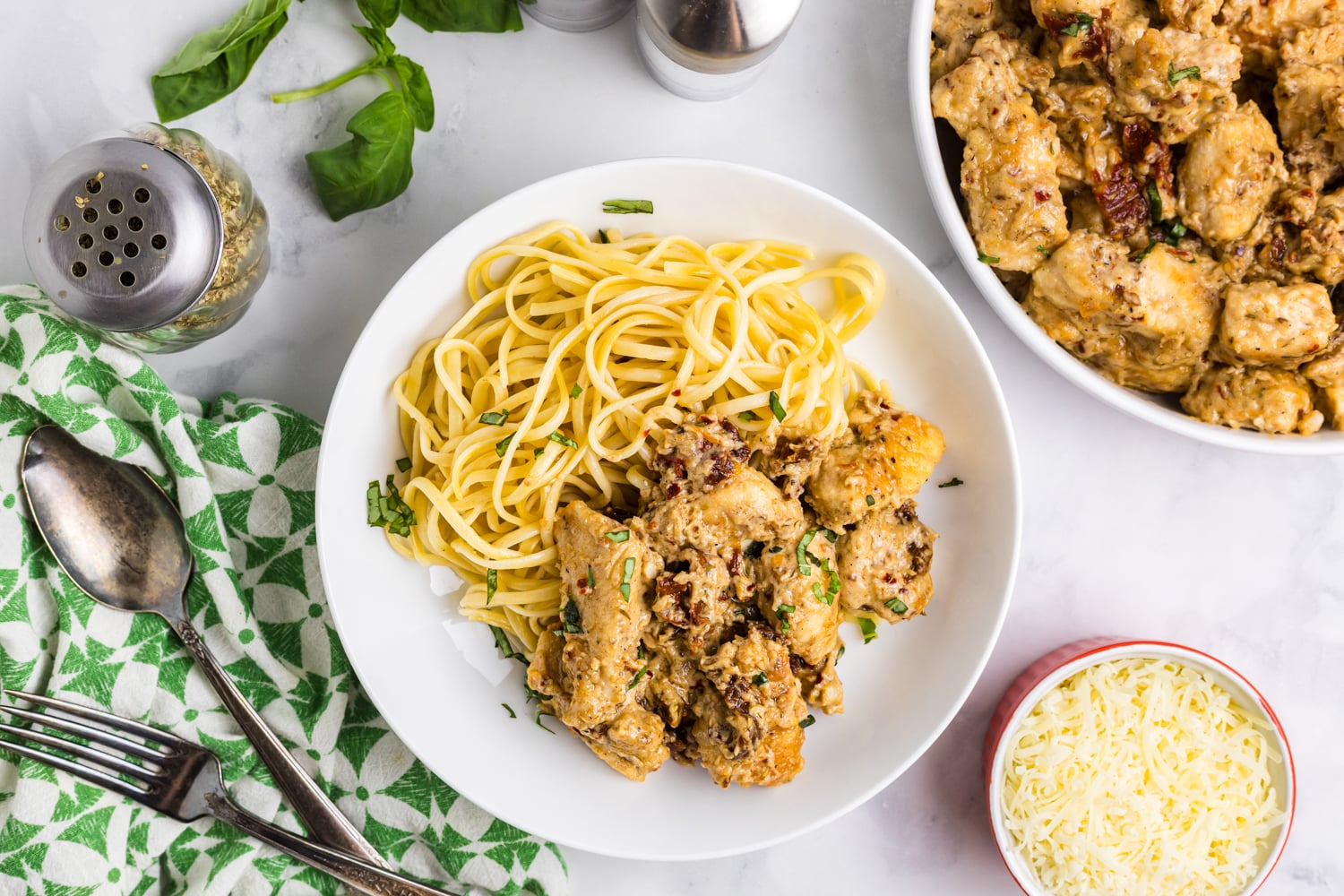 Overhead view of a bowl of pasta topped with creamy sun-dried tomato chicken, garnished with herbs and surrounded by utensils, cheese, basil leaves, and seasonings on a light countertop.