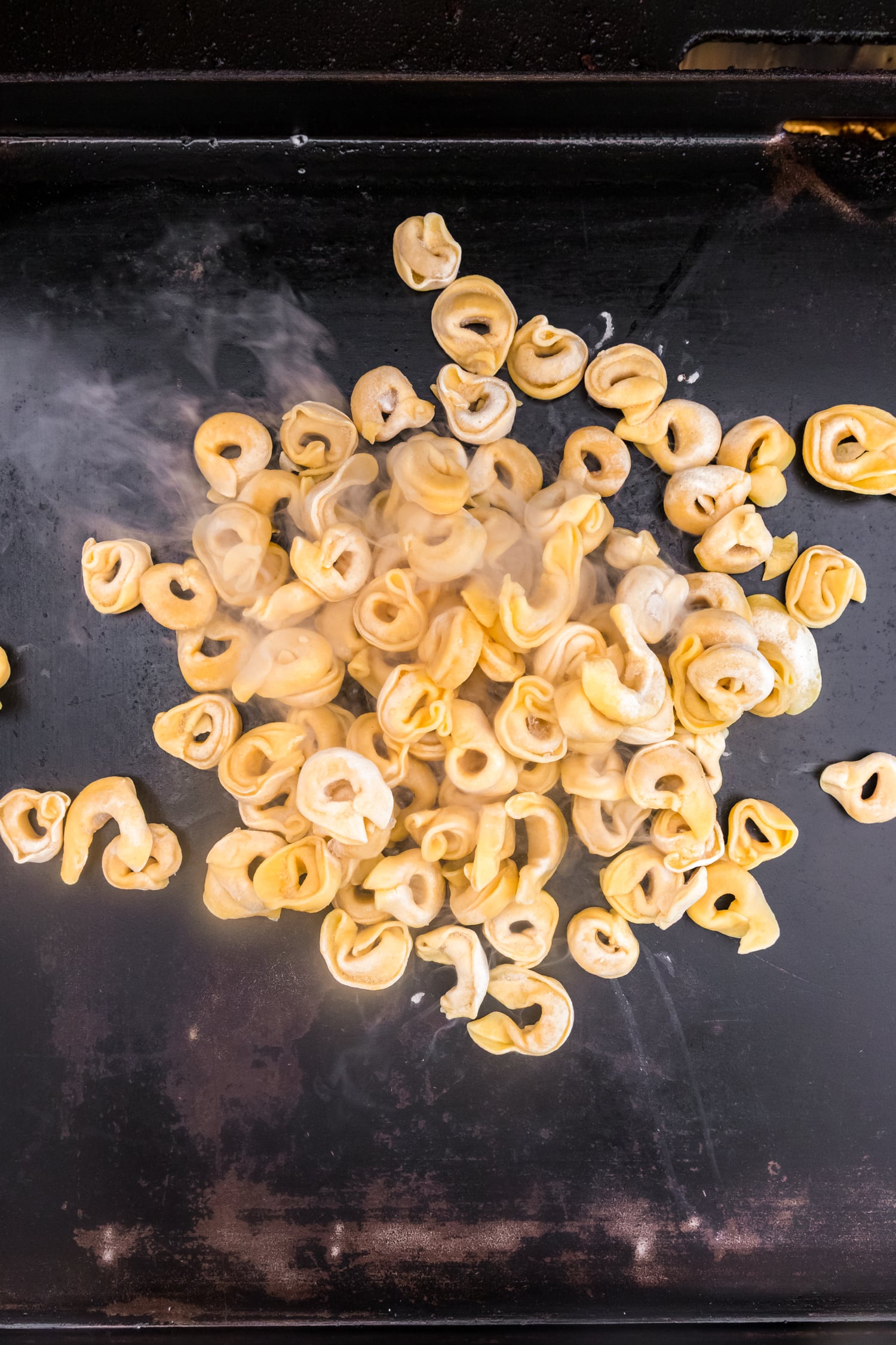 Cheese tortellini cooking on a hot flat-top griddle with steam rising.