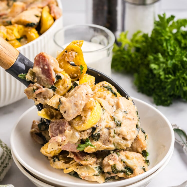 Serving spoon lifting creamy chicken bacon tortellini with spinach into a white bowl.