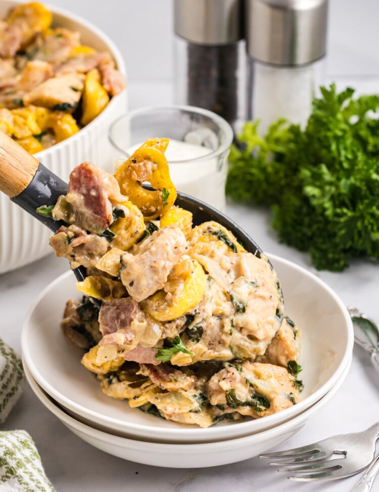Serving spoon lifting creamy chicken bacon tortellini with spinach into a white bowl.