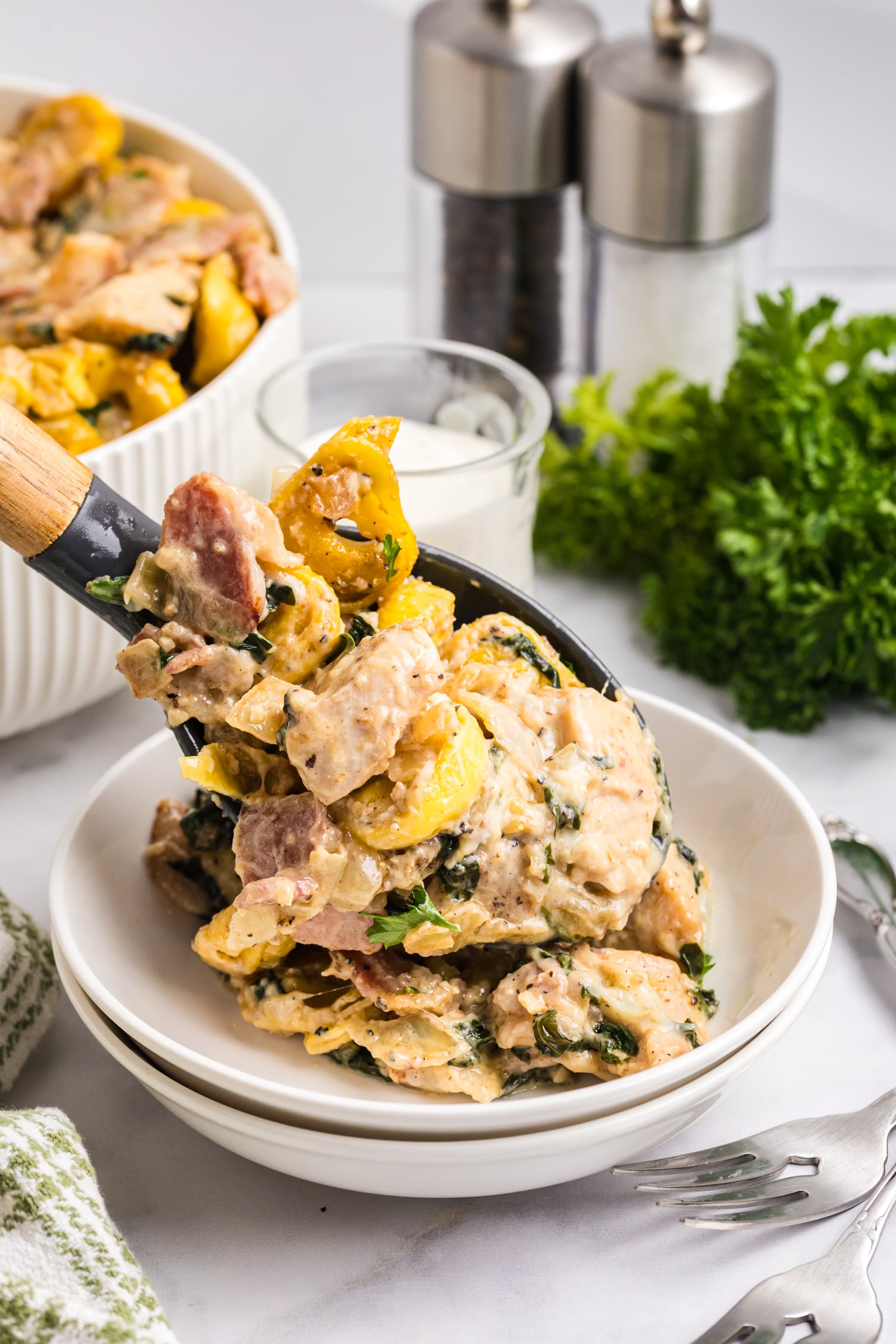 Serving spoon lifting creamy chicken bacon tortellini with spinach into a white bowl.