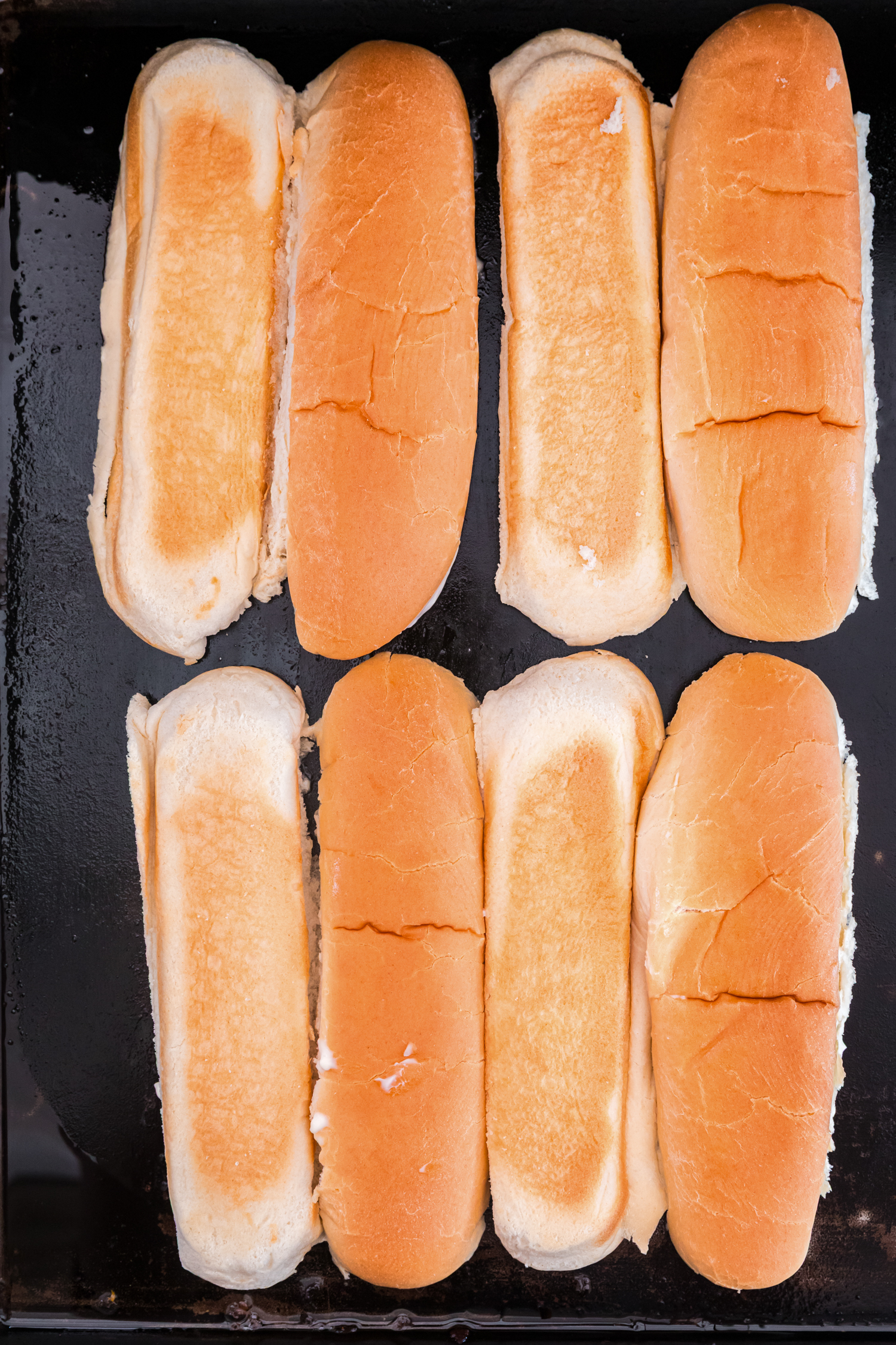 Several split hoagie rolls arranged on a flat griddle, lightly toasted on the inside.