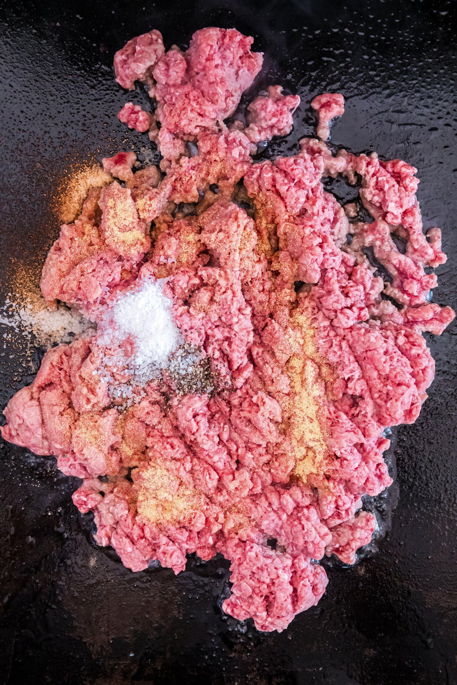 Ground beef cooking on a flat-top griddle with salt, pepper, and seasoning sprinkled on top.