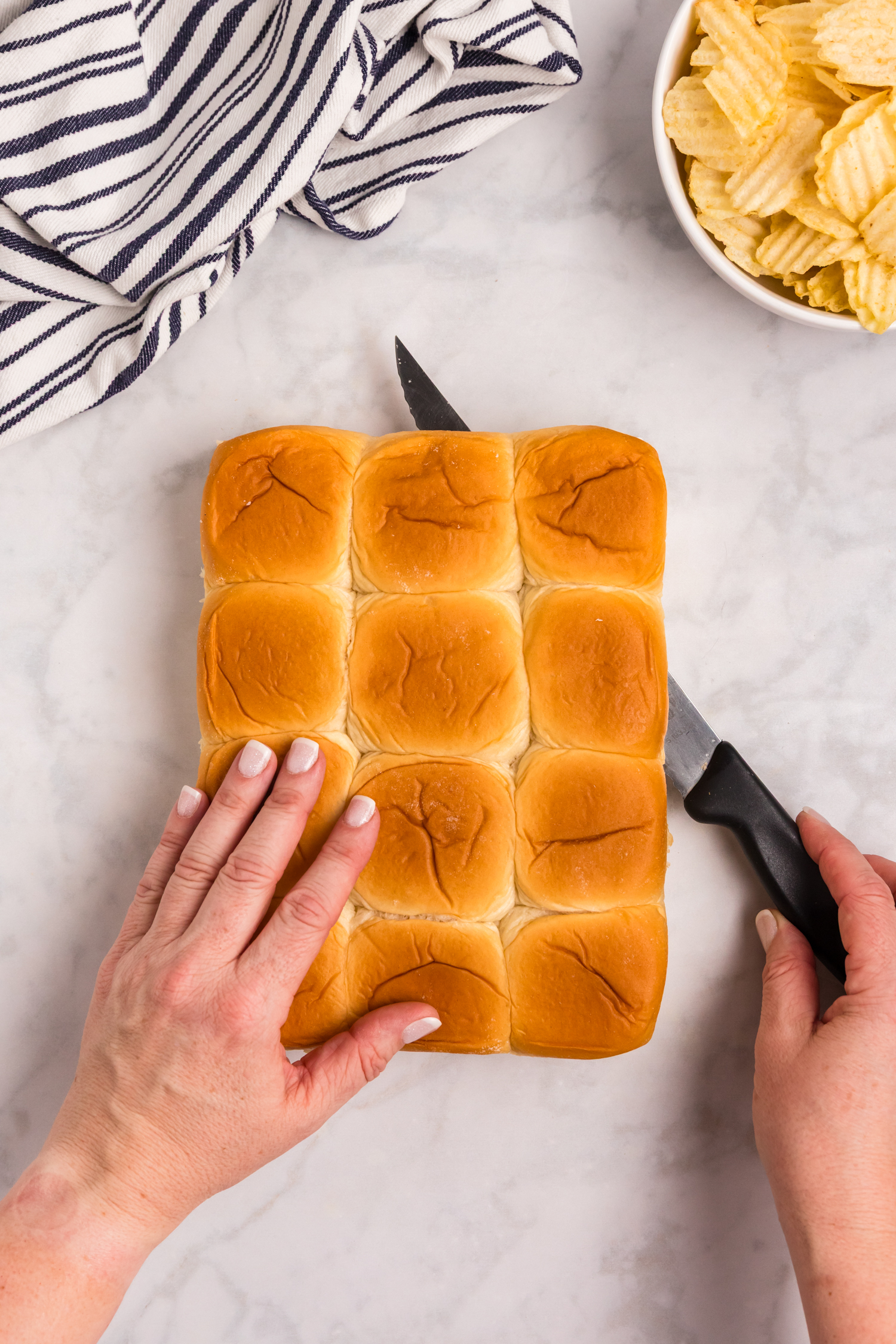 Hands slicing a sheet of soft slider buns with a knife on a marble surface, with chips and a kitchen towel nearby.