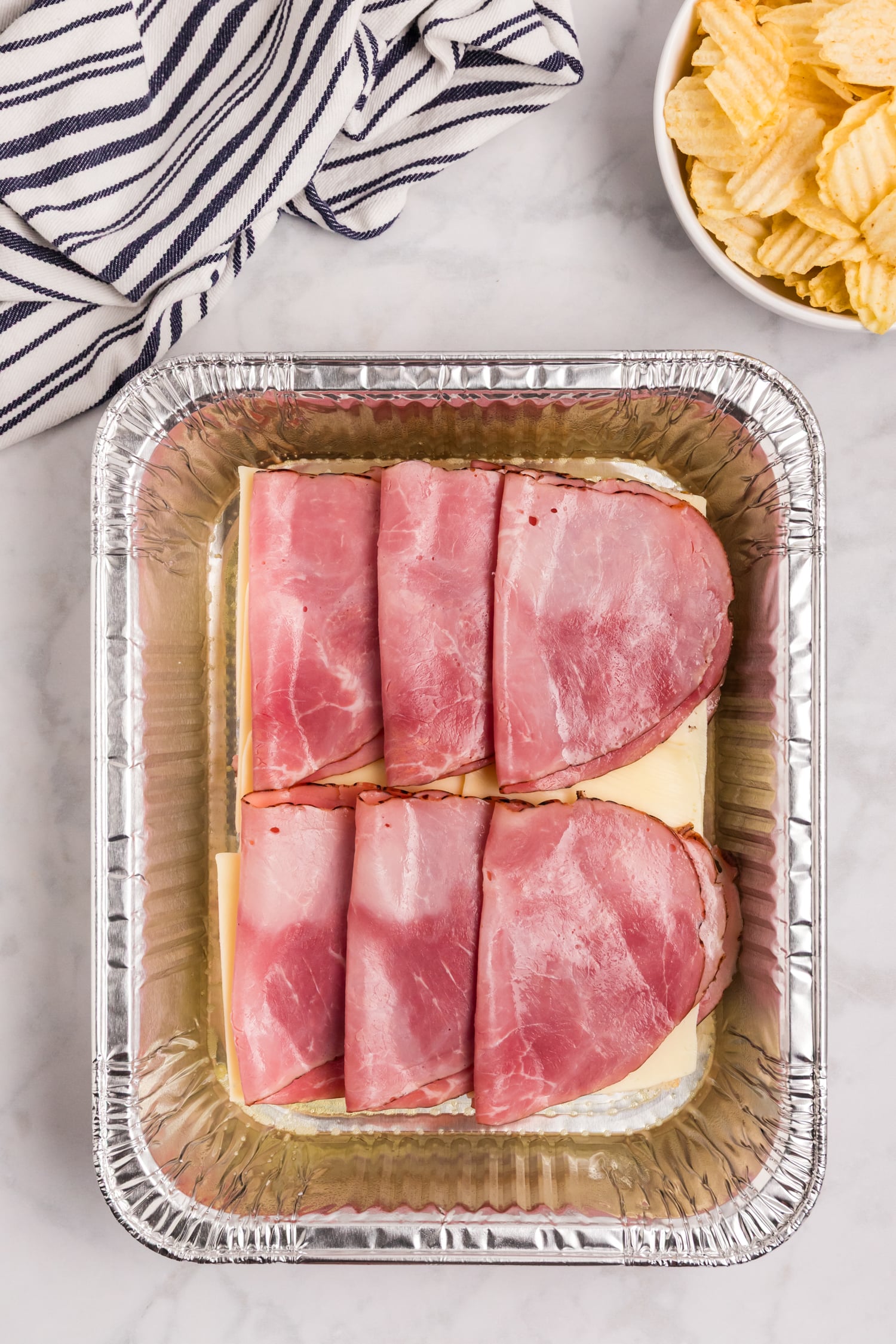 Bottom layer of slider buns topped with sliced cheese and folded ham inside a foil baking pan.