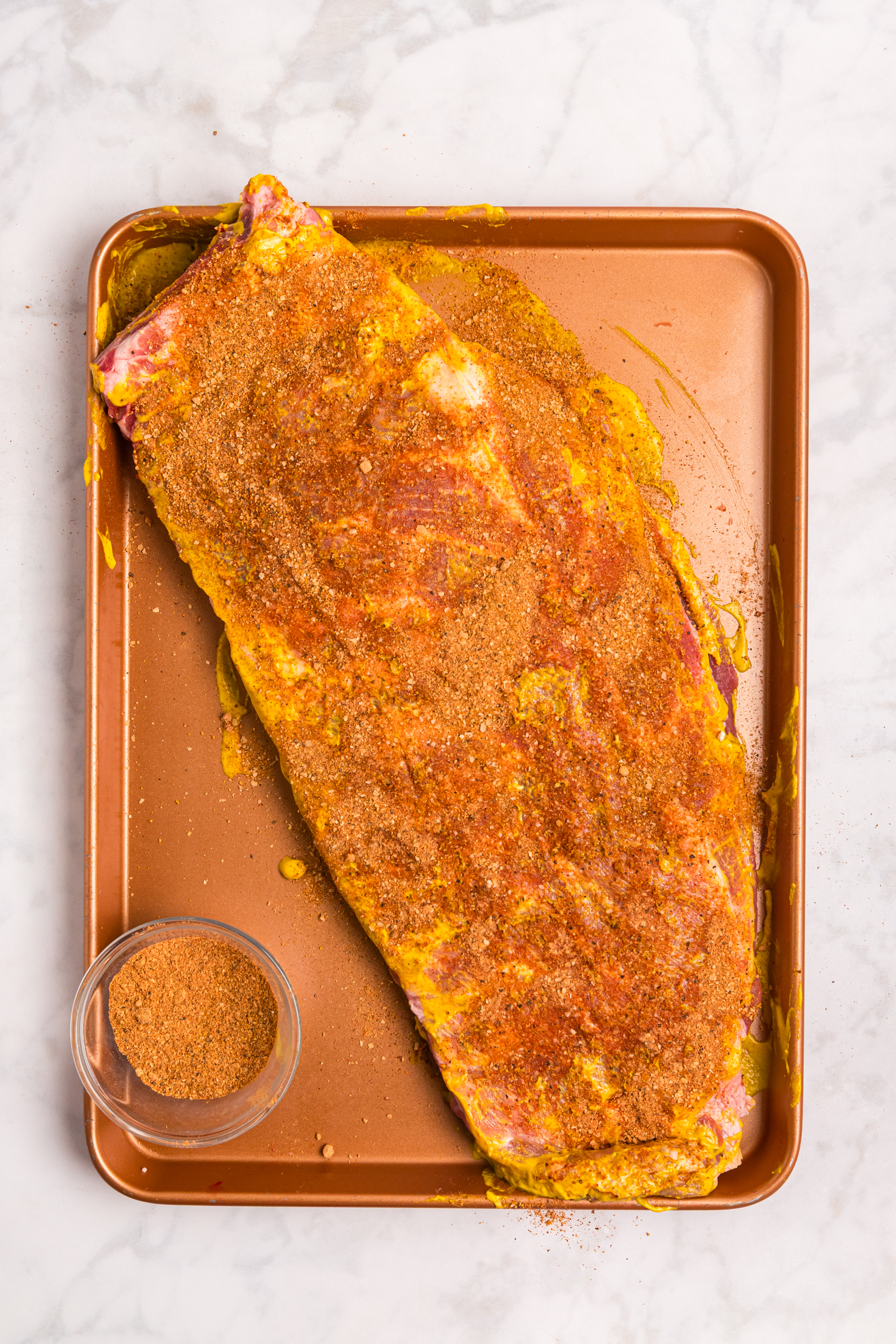 Raw pork ribs coated with mustard and generously covered in dry rub seasoning on a sheet pan.
