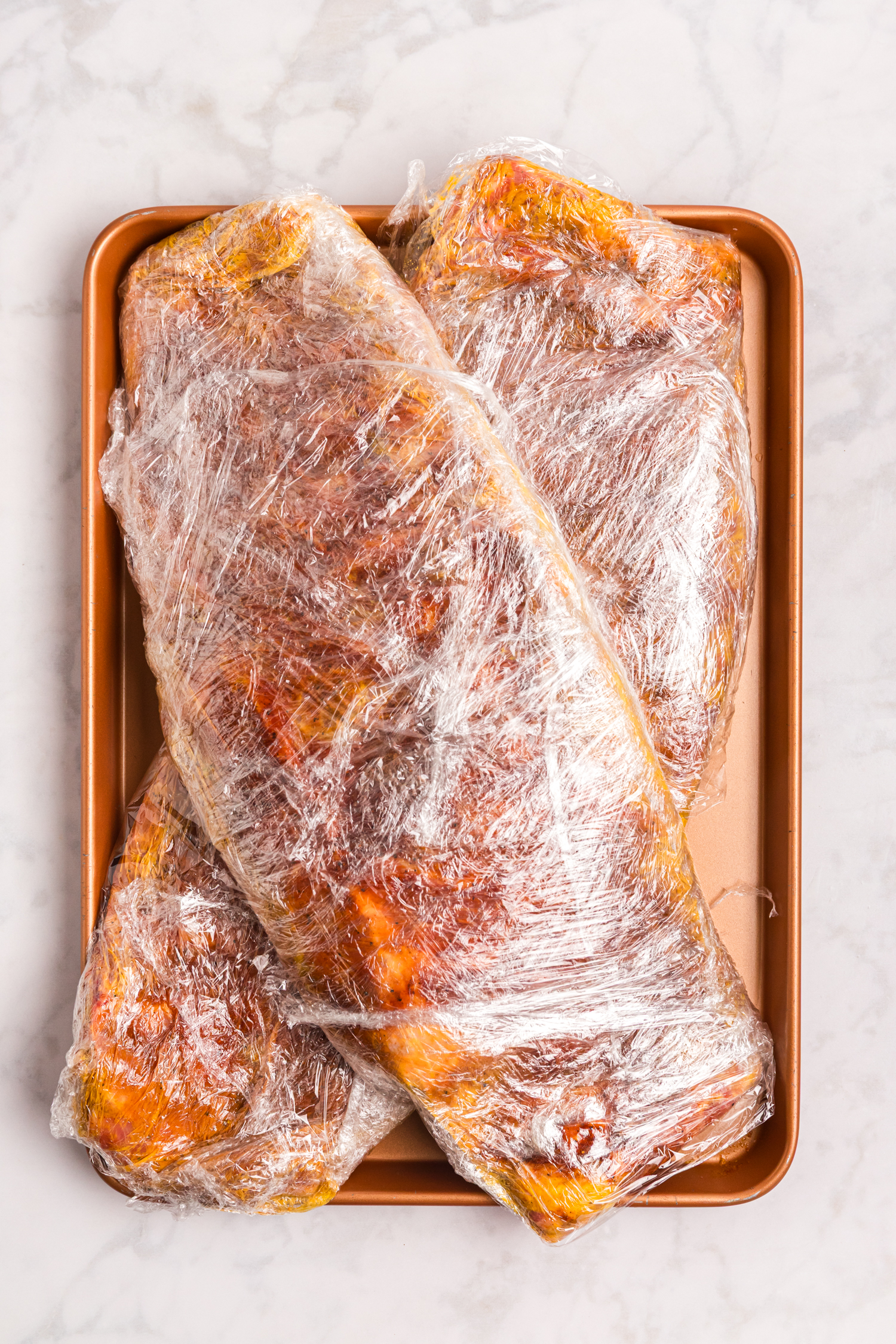 Seasoned racks of ribs tightly wrapped in plastic wrap resting on a baking sheet for marinating.