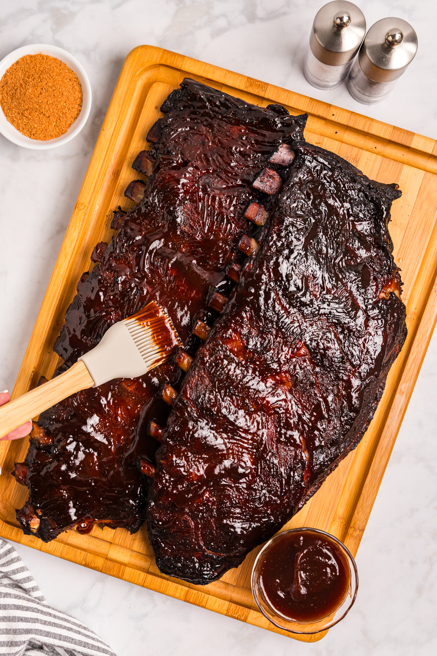 Cooked pork ribs on a wooden board being brushed with barbecue sauce, with seasoning and sauce nearby.