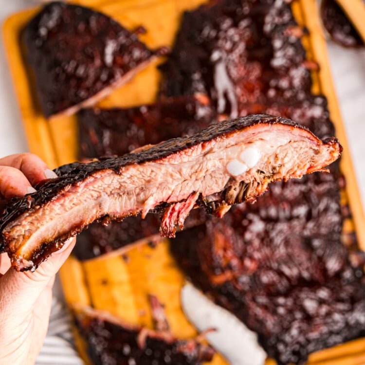 Close-up of a hand holding a sliced smoked rib showing a juicy interior and bark, with more ribs in the background.