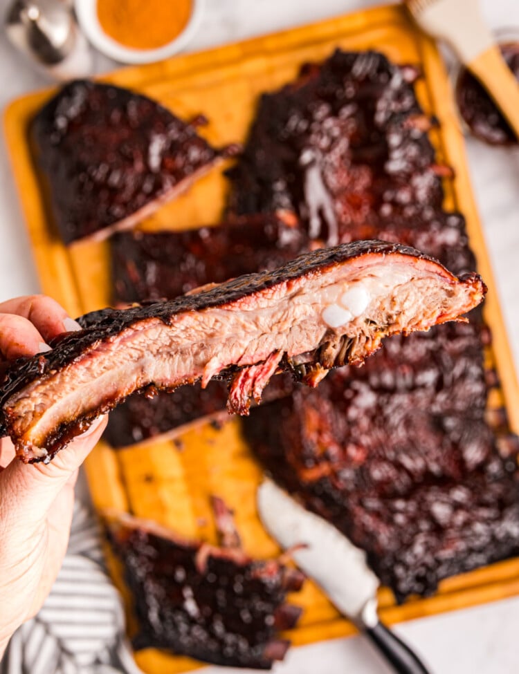Close-up of a hand holding a sliced smoked rib showing a juicy interior and bark, with more ribs in the background.