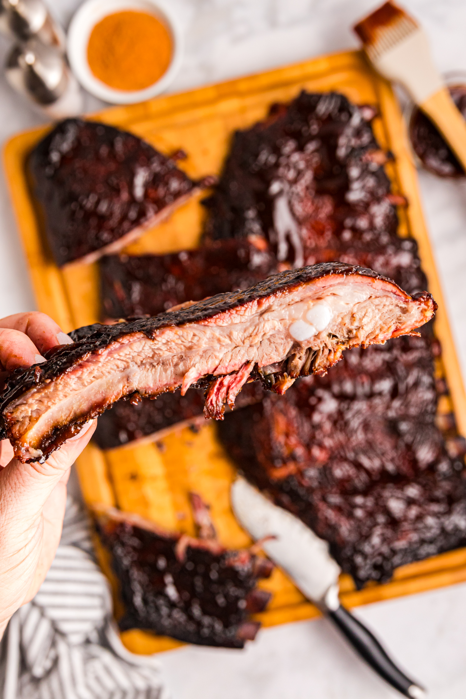 Close-up of a hand holding a sliced smoked rib showing a juicy interior and bark, with more ribs in the background.