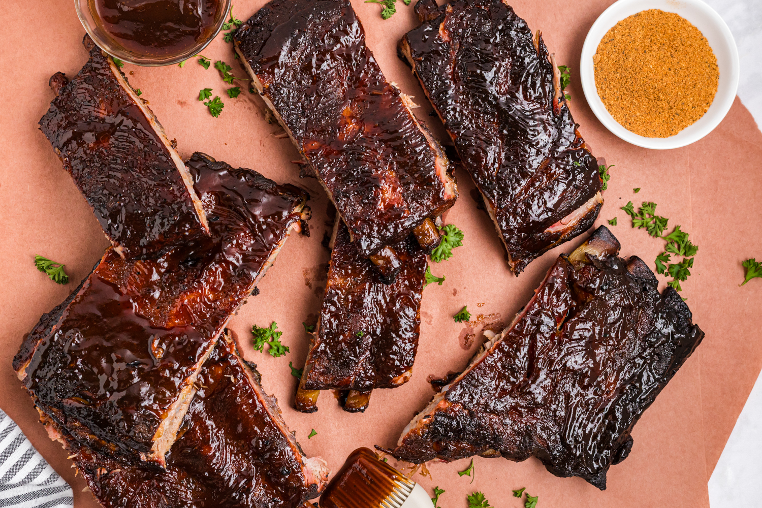 Close-up of sliced smoked pork ribs coated in glossy barbecue sauce, arranged on parchment paper with a bowl of dry rub and fresh herbs.