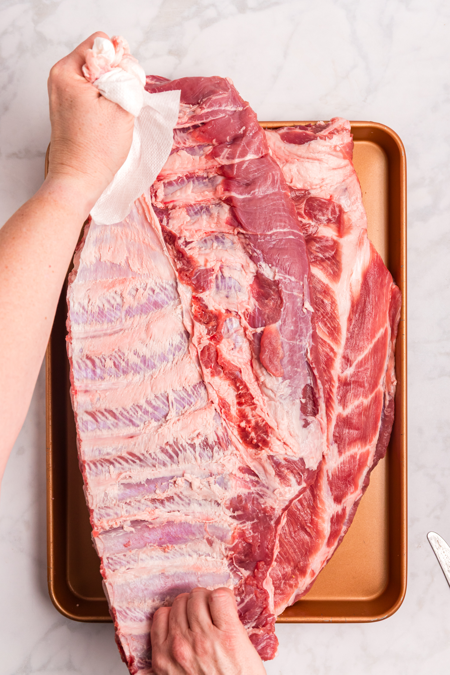 Hands pulling the membrane off the back of a raw rack of pork ribs on a baking sheet.