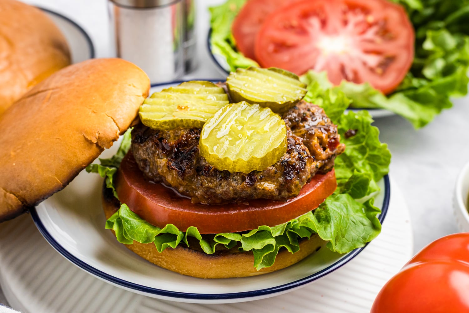 A grilled cheeseburger on a bun topped with pickle slices, lettuce, and a thick tomato slice, with the top bun slightly lifted.