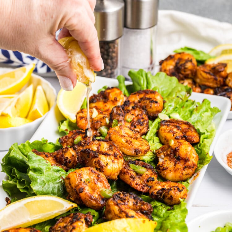 Hand squeezing lemon juice over grilled shrimp served on lettuce with lemon wedges and green onions.