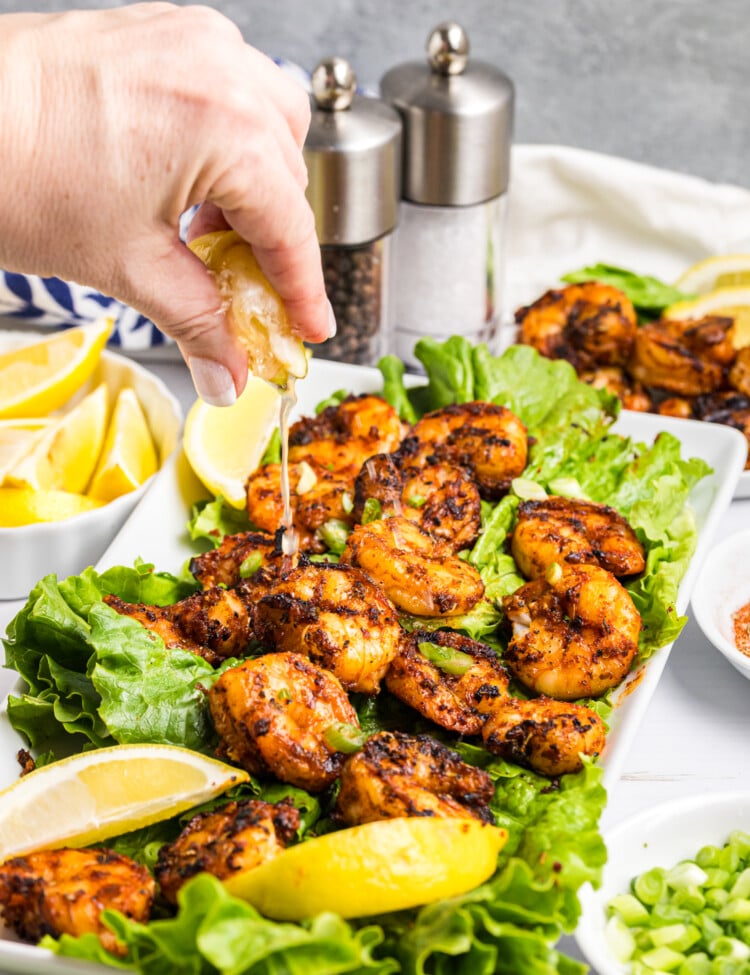Hand squeezing lemon juice over grilled shrimp served on lettuce with lemon wedges and green onions.